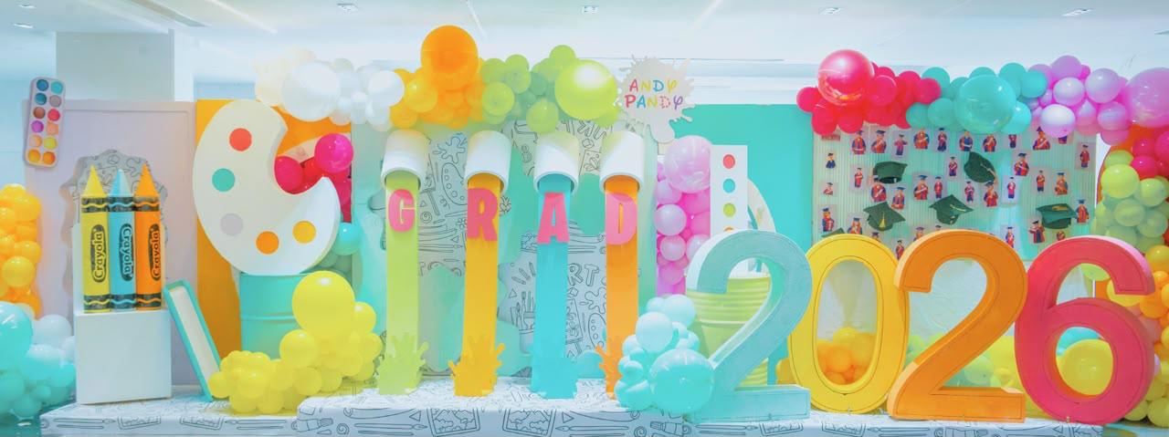 Colorful GRAD 2026 stage decoration at Andy Pandy graduation ceremony, with balloon arches and giant crayon and paint palette props
