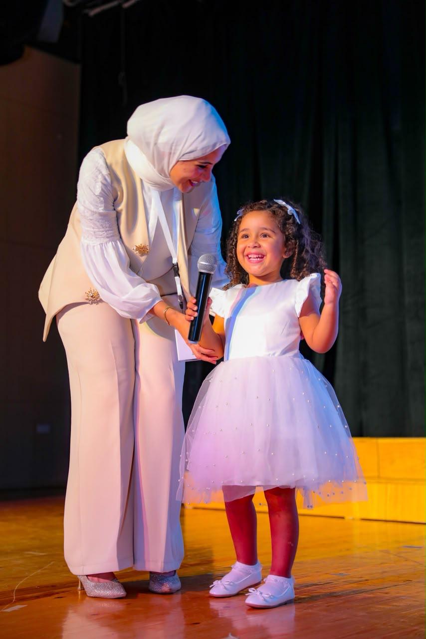 Andy Pandy teacher handing the microphone to a joyful little girl in a white dress on stage